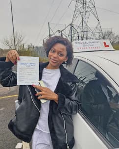 Smiling woman holds a practical driving test pass certificate next to a white car.