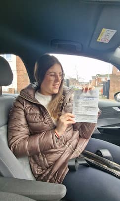 A young woman in a car holding her practical driving test pass certificate.