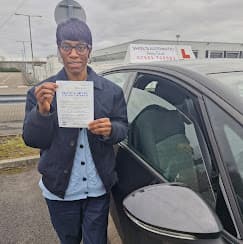 A woman proudly holds her practical driving test pass certificate next to a learner car.