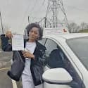 Smiling woman holds a driving test pass certificate next to a white learner driver car.