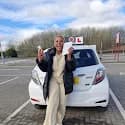 Smiling woman holds her driving test pass certificate and license in front of white car.